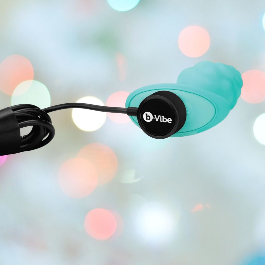 A B-Vibe Texture Vibrating Butt Plug Bump - Mint (Small), made of body-safe silicone, is charging with a black magnetic USB charger against a backdrop of blurred pastel lights.
