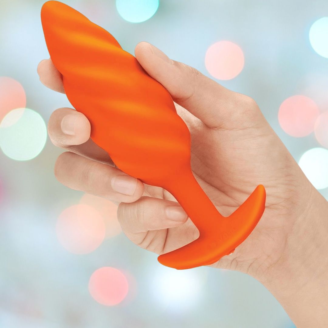 A hand holds the B-Vibe Texture Vibrating Butt Plug Swirl in orange (medium), a body-safe silicone toy from B-Vibe, against a blurred background of colorful lights—ideal for adventurous anal play.