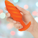A hand holds the B-Vibe Texture Vibrating Butt Plug Swirl in orange (medium), a body-safe silicone toy from B-Vibe, against a blurred background of colorful lights—ideal for adventurous anal play.