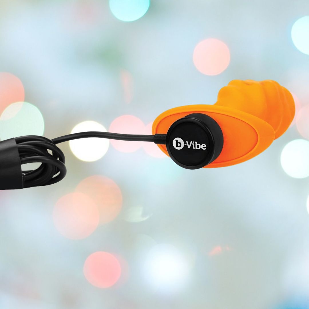 The B-Vibe Texture Vibrating Butt Plug Swirl - Orange (Medium) features ridged body-safe silicone and a prominent black "b-Vibe" logo, shown charging against a colorful bokeh background—ideal for anal play lovers.