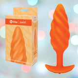 The B-Vibe Texture Vibrating Butt Plug Swirl - Orange (Medium) by B-Vibe, featuring a swirl-textured silicone design, is displayed beside its box against a soft, colorful background with circular light spots.