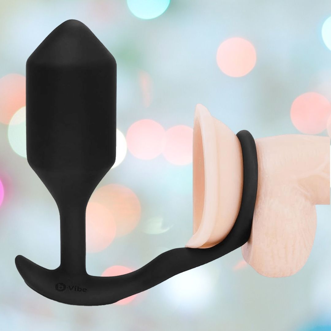 The B-Vibe Vibrating Cock Ring - Snug & Tug (M), a black silicone butt plug and penis ring by B-Vibe, is shown on a realistic penis model with a blurred pastel background and soft circular light spots.