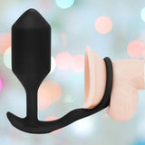 The B-Vibe Vibrating Cock Ring - Snug & Tug (M), a black silicone butt plug and penis ring by B-Vibe, is shown on a realistic penis model with a blurred pastel background and soft circular light spots.