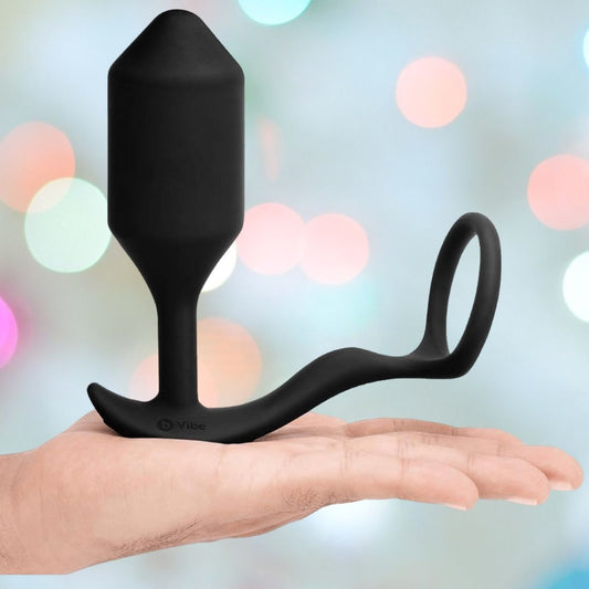 A hand holds the B-Vibe Vibrating Cock Ring - Snug & Tug (M) by B-Vibe, featuring black silicone, a curved base, and a finger ring, set against a blurred pastel background with circular lights. 1080