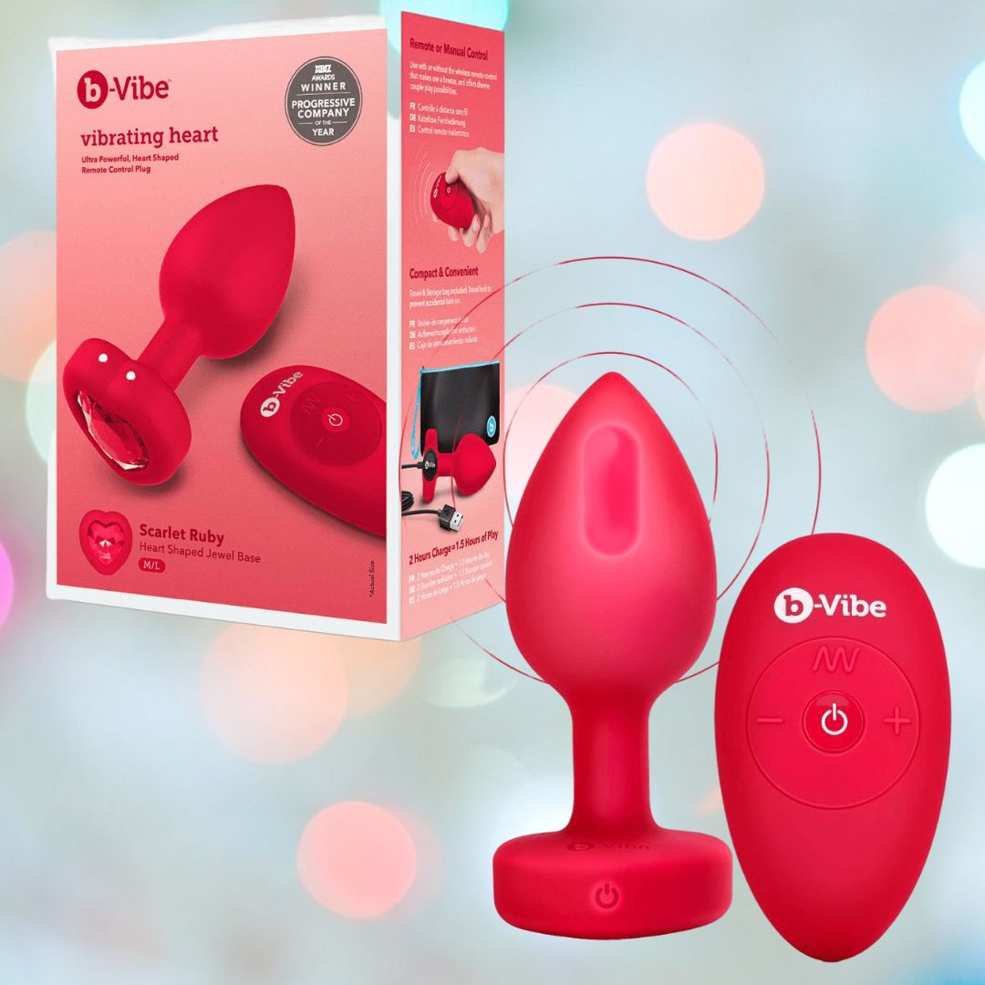 The B-Vibe Vibrating Heart Plug Medium/Large in Scarlet Ruby is shown beside its packaging, featuring remote control and highlighting the product name "B-Vibe Vibrating Heart Plug" by B-Vibe on a light gradient background.