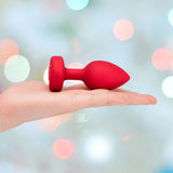 A hand holds the B-Vibe Vibrating Heart Plug Medium/Large in Scarlet Ruby by B-Vibe, set against a softly blurred pastel background, evoking playful and romantic vibes.