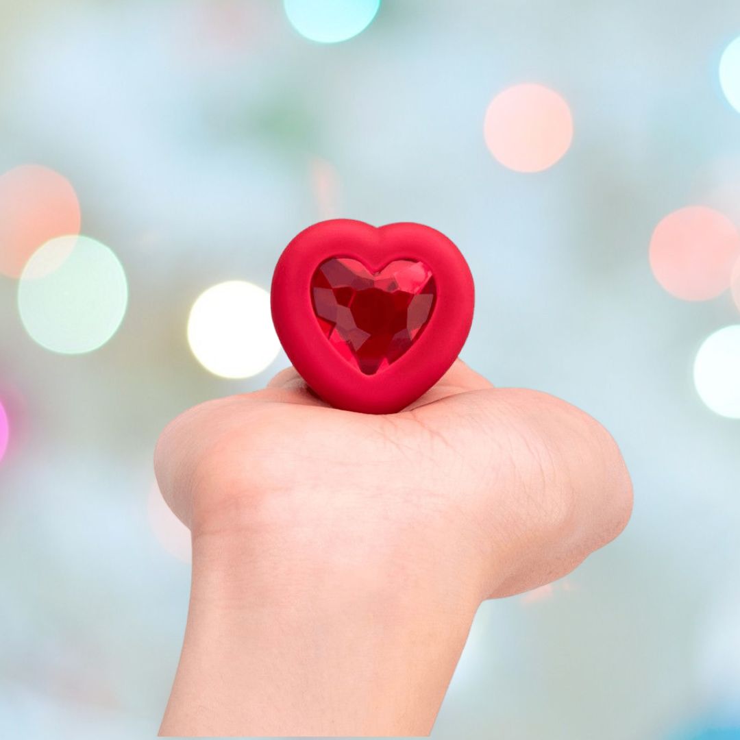 A hand holds the B-Vibe Vibrating Heart Plug Medium/Large – Scarlet Ruby by B-Vibe, its red heart shape and gem-like center gleaming against a softly blurred bokeh background, evoking playful possibilities.