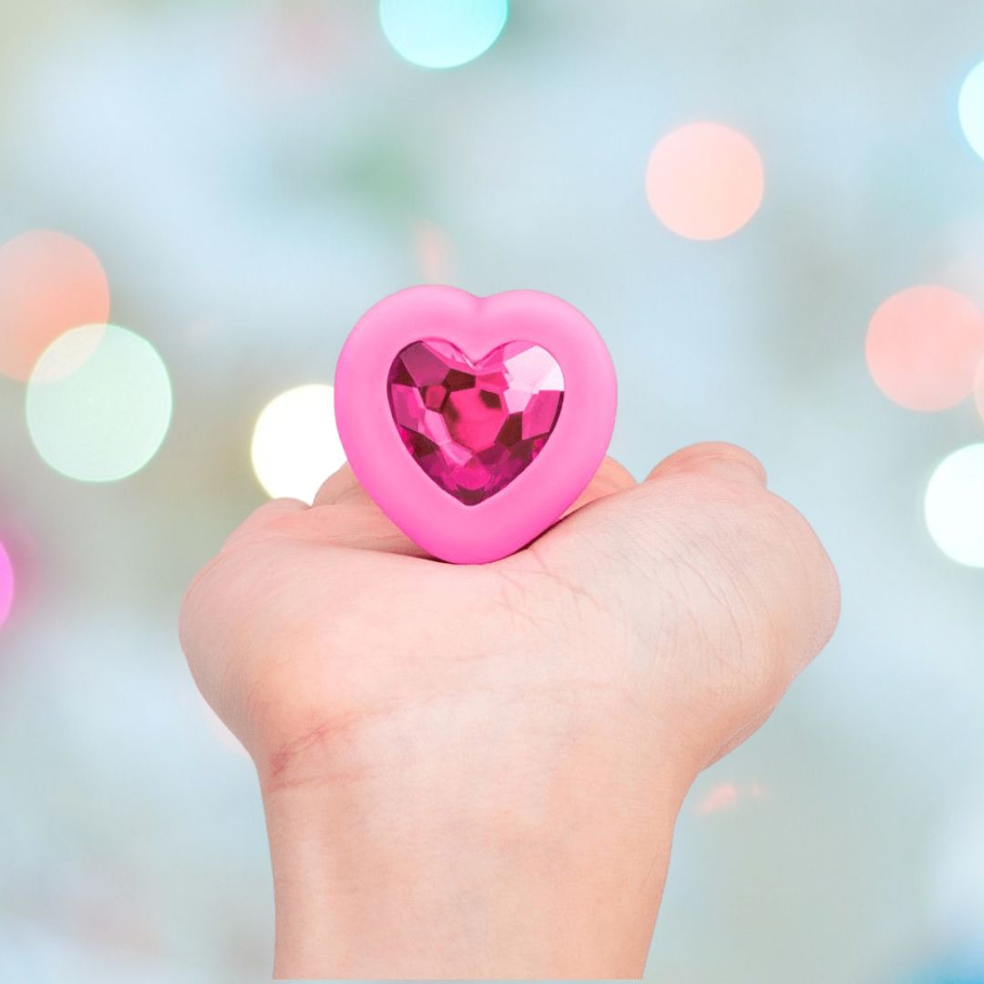 A hand holds the B-Vibe Vibrating Heart Plug Small/Medium - Pink Topaz, featuring a gem-like center, against a softly lit pastel background, suggesting playful jeweled stimulation.