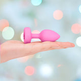 The B-Vibe Vibrating Heart Plug Small/Medium - Pink Topaz is displayed on an outstretched hand against a colorful, bokeh-style background—bringing stylish comfort to anal play for beginners and enthusiasts alike.