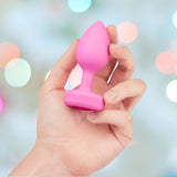A hand holds the B-Vibe Vibrating Heart Plug Small/Medium in Pink Topaz, featuring a power button at the base—perfect for anal play—against a blurred, colorful background.