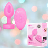 A B-Vibe Vibrating Heart Plug Small/Medium - Pink Topaz is shown next to its branded packaging on a pastel background, highlighting its wireless remote and stylish design for enhanced anal play.