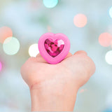 A hand holds the B-Vibe Vibrating Heart Plug Small/Medium - Pink Topaz, featuring a gem-like center, against a softly lit pastel background, suggesting playful jeweled stimulation.