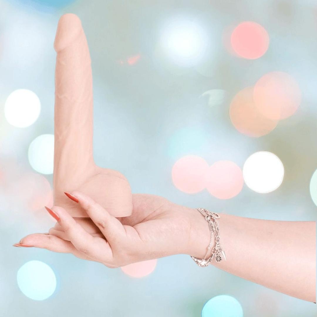A hand with red nails and a silver bracelet holds the BMS Naked Addiction Thursting Dong - Vanilla 8.5" upright against a softly blurred pastel backdrop with circular light spots.