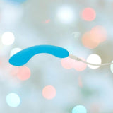 The BMS The Mini Swan Wand Glow in the Dark - Blue is a curved vibrator with a white cord, shown against a blurred background of pastel lights.