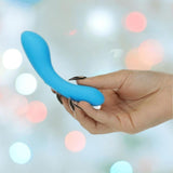 A hand with long black nails holds the BMS The Mini Swan Wand Glow in the Dark - Blue by BMS against a softly blurred pastel-lit background.