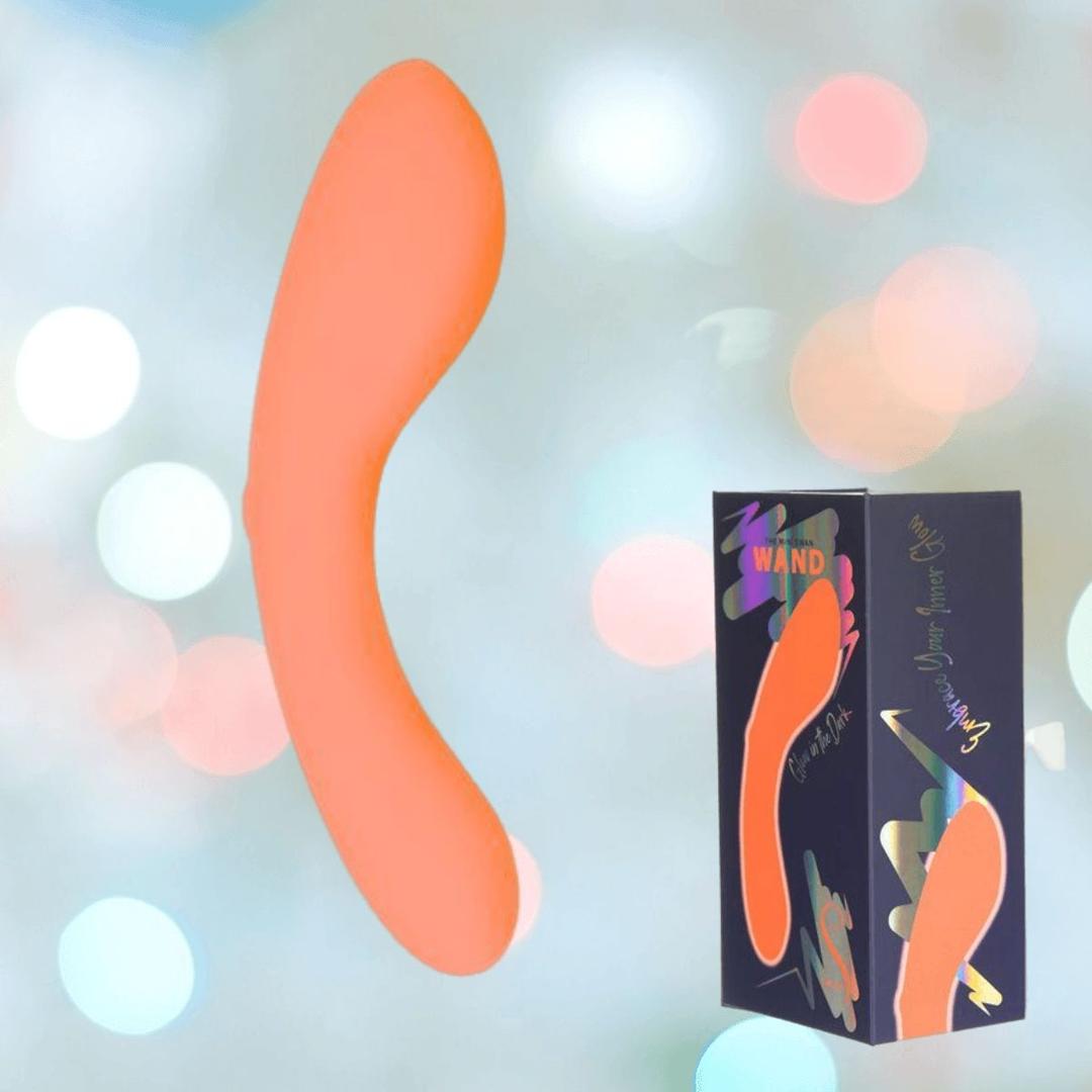 The BMS The Mini Swan Wand Glow in the Dark-Orange, a curved orange vibrator, floats next to a dark blue box with its shape and colorful text. A soft light background features out-of-focus pastel circles.