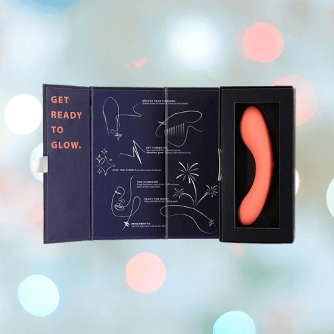 A BMS The Mini Swan Wand Glow in the Dark-Orange vibrator, shown in a black case and open packaging with instructions and "GET READY TO GLOW" printed inside. The pastel-lit background highlights its playful, vibrant design.