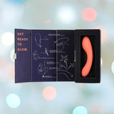 A BMS The Mini Swan Wand Glow in the Dark-Orange vibrator, shown in a black case and open packaging with instructions and "GET READY TO GLOW" printed inside. The pastel-lit background highlights its playful, vibrant design.