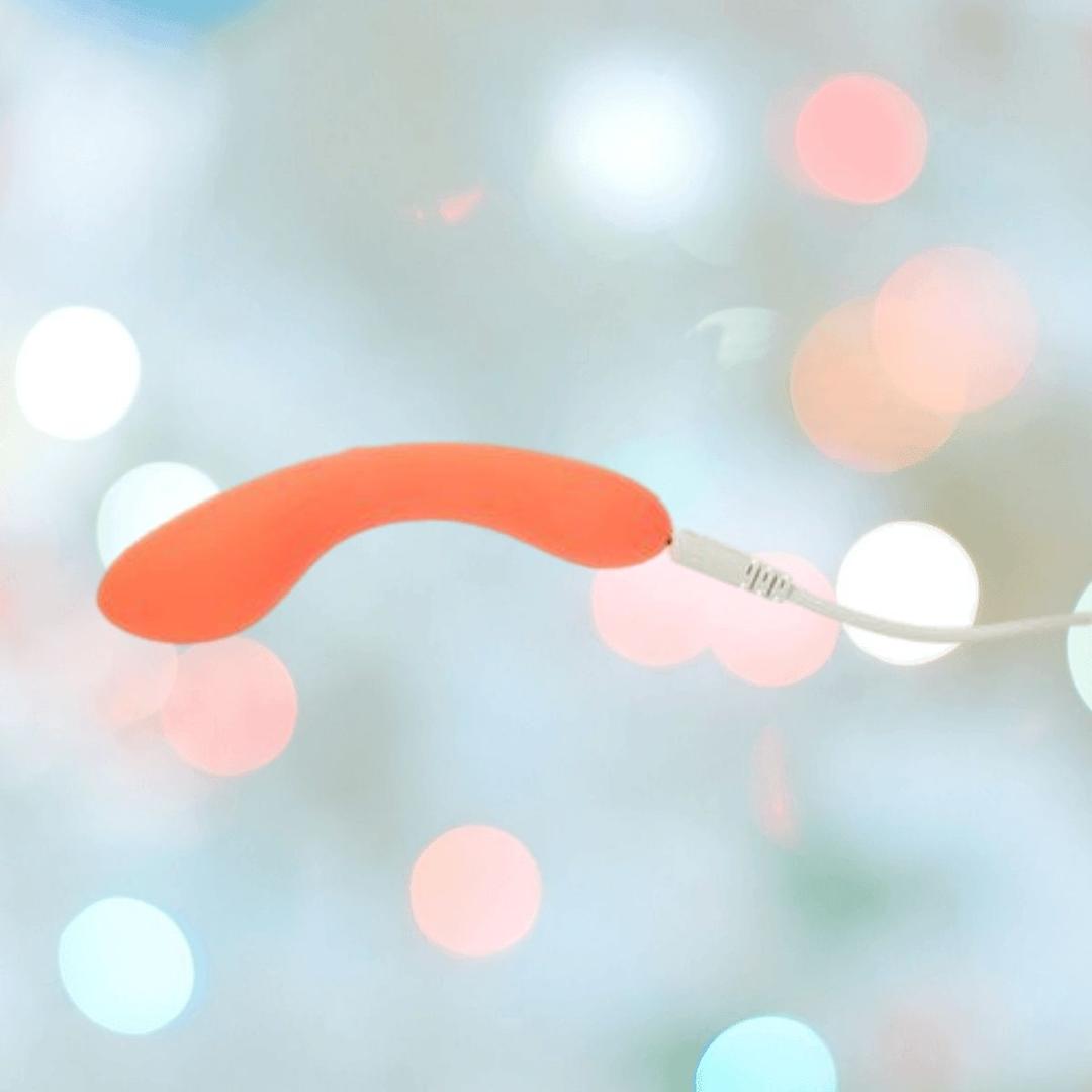 The BMS The Mini Swan Wand Glow in the Dark-Orange vibrator glows as it connects to its white charging cable, with a blurred pastel bokeh background highlighting the vibrant product by BMS.