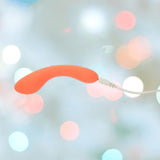 The BMS The Mini Swan Wand Glow in the Dark-Orange vibrator glows as it connects to its white charging cable, with a blurred pastel bokeh background highlighting the vibrant product by BMS.