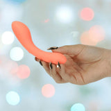 A hand with long, metallic nails holds the BMS The Mini Swan Wand Glow in the Dark-Orange vibrator against a blurred background with soft white and pink lights.