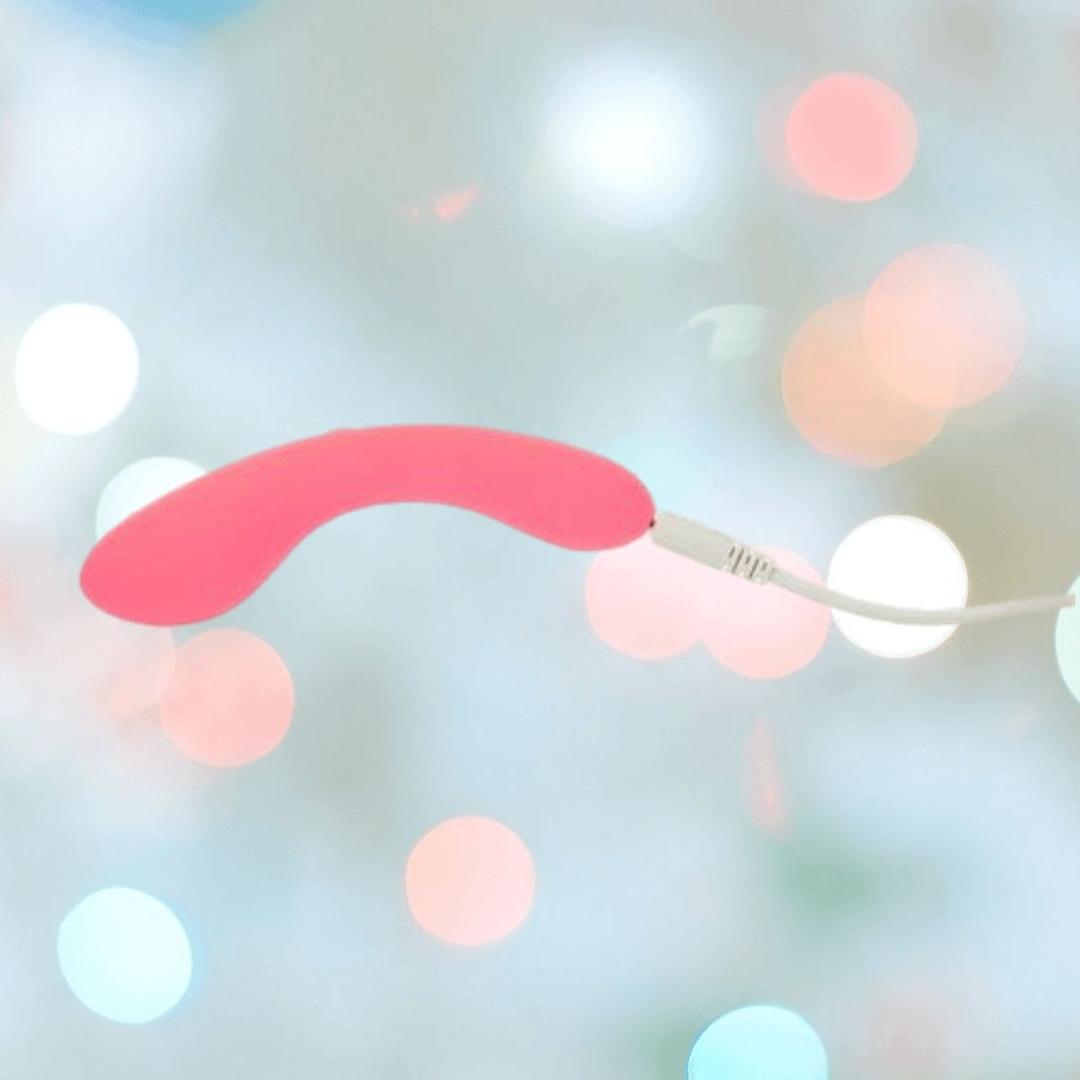 The BMS The Mini Swan Wand Glow in the Dark - Pink by bms is shown connected to a white charging cable, set against a blurred background with pastel bokeh lights.