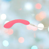The BMS The Mini Swan Wand Glow in the Dark - Pink by bms is shown connected to a white charging cable, set against a blurred background with pastel bokeh lights.