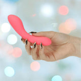A hand with painted nails holds the bms The Mini Swan Wand Glow in the Dark - Pink, its curved silicone shape glowing softly against a colorful, blurred background.