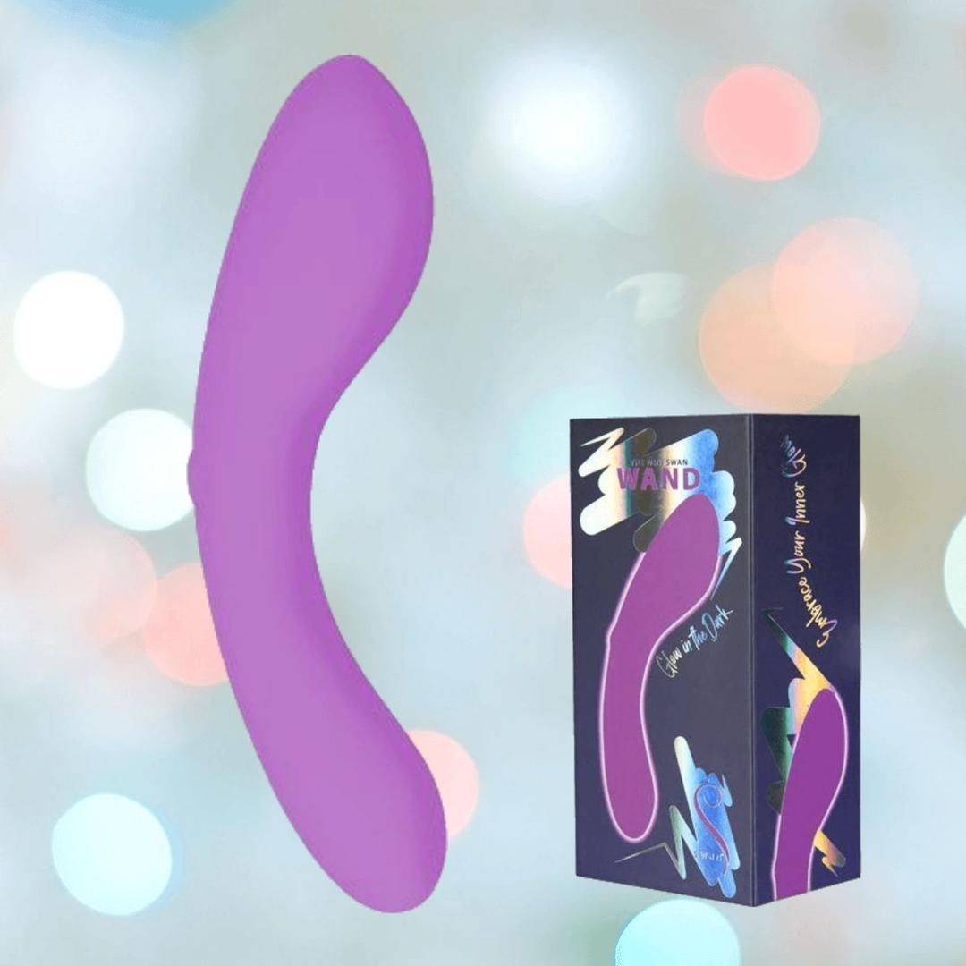The BMS The Mini Swan Wand Glow in the Dark-Purple, a curved and compact vibrator, floats near its branded packaging with matching imagery and vibrant abstract designs, set against a softly blurred pastel background.