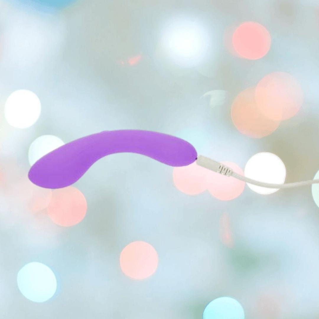 The BMS The Mini Swan Wand Glow in the Dark-Purple, a compact vibrator by BMS, is shown connected to a white charging cable against a backdrop of blurred, softly colored lights.