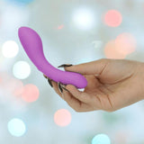 A hand with long, black nails holds the BMS The Mini Swan Wand Glow in the Dark-Purple, a compact curved vibrator, against a soft background with colorful bokeh lights.