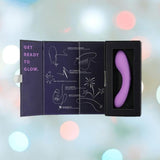 A BMS The Mini Swan Wand Glow in the Dark-Purple vibrator rests in its open black box, revealing playful line art and instructions inside. The box reads "GET READY TO GLOW," set against a softly blurred background of pastel lights.
