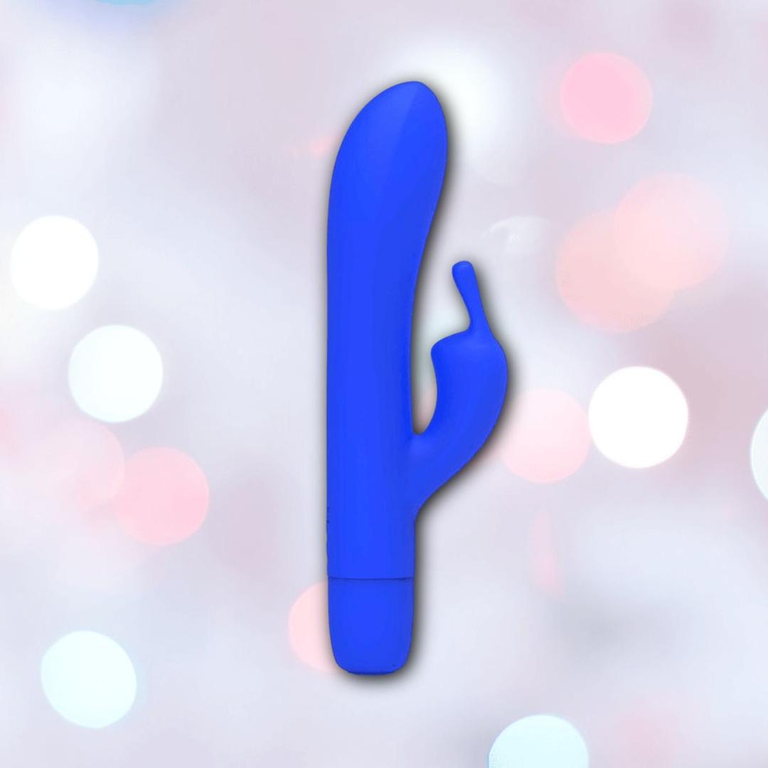 The B Swish Bwild Classic Infinite Bunny - Pacific Blue by Condomania.com, a blue silicone rabbit vibrator with a curved shaft and smaller arm, is shown against a soft pastel background with circular light bokeh.