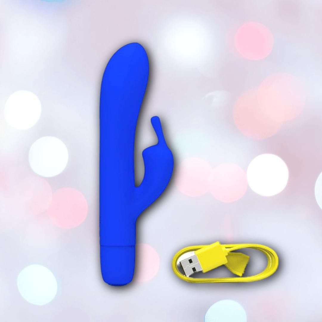 The B Swish Bwild Classic Infinite Bunny - Pacific Blue from Condomania.com, a blue silicone rabbit vibrator with a curved shaft and small extension, is shown beside a yellow USB charging cable on a soft pink and white bokeh background.