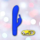 The B Swish Bwild Classic Infinite Bunny - Pacific Blue from Condomania.com, a blue silicone rabbit vibrator with a curved shaft and small extension, is shown beside a yellow USB charging cable on a soft pink and white bokeh background.