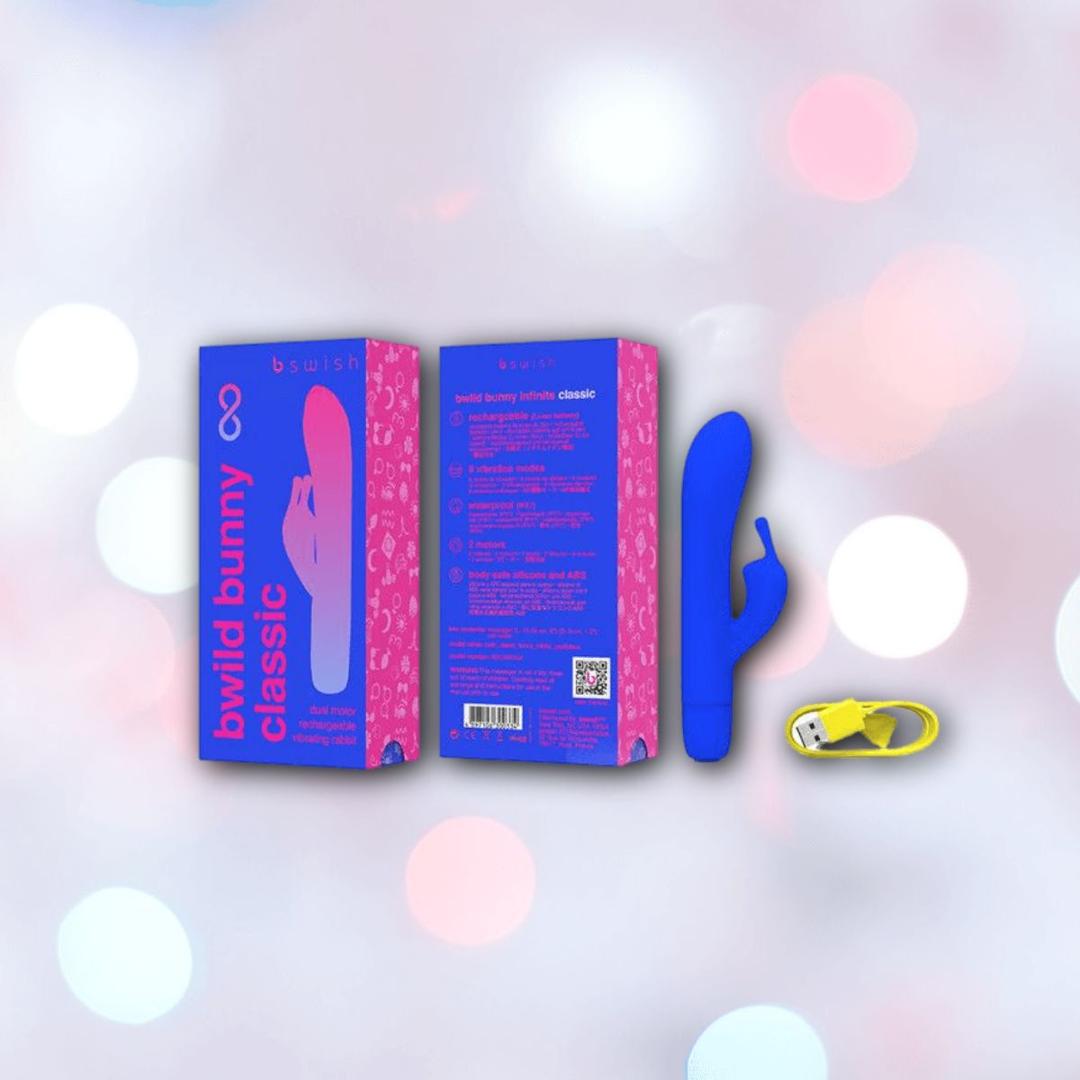 A Pacific blue B Swish Bwild Classic Infinite Bunny vibrator from Condomania.com is shown beside its blue and pink box and a small yellow battery, all set against a soft, bokeh background.