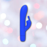 The B Swish Bwild Classic Infinite Bunny - Pacific Blue by Condomania.com, a blue silicone rabbit vibrator with a curved shaft and smaller arm, is shown against a soft pastel background with circular light bokeh.