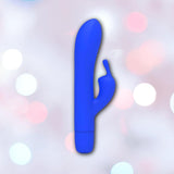 The B Swish Limited Edition Bwild Bunny - Pacific Blue (Dented Box) features a curved shaft and a smaller silicone clitoral stimulator, shown on a pastel bokeh background.