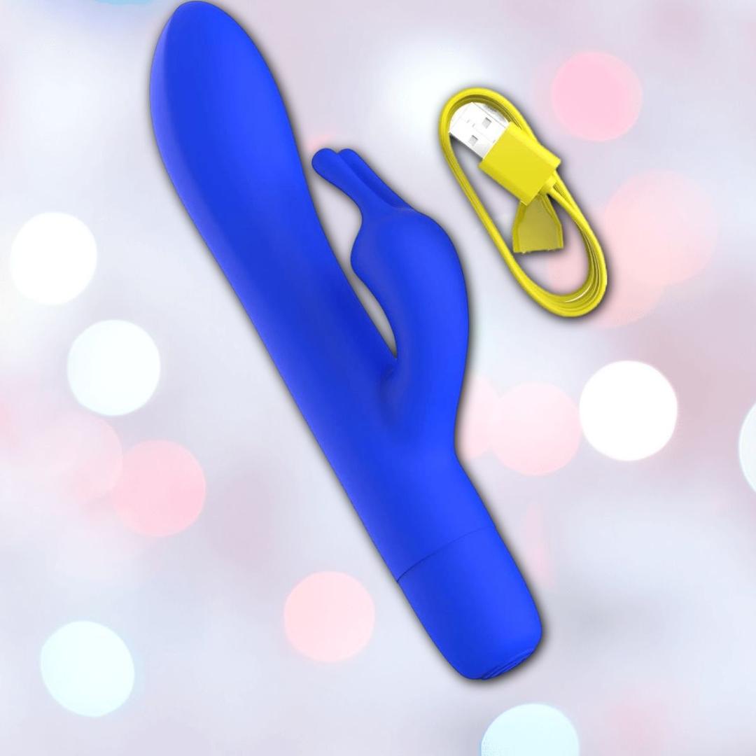A B Swish Limited Edition Bwild Bunny - Pacific Blue (Dented Box) silicone stimulator with a curved shaft and small arm is pictured next to a yellow USB charging cable on a pastel, softly blurred background.