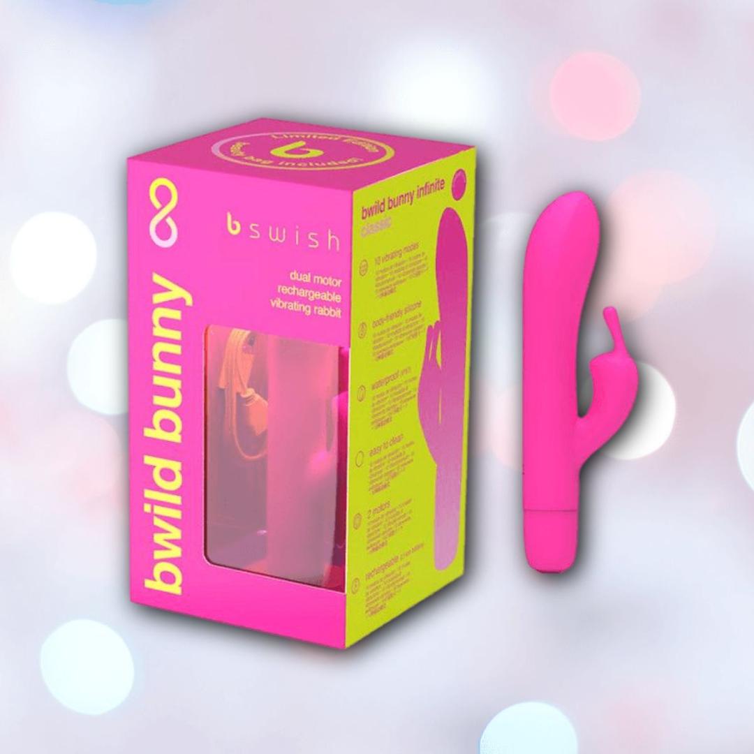 The B Swish Limited Edition Bwild Infinite Bunny - Sunset Pink, a waterproof silicone vibrator with dual motors, is displayed next to its eye-catching yellow and pink box against a backdrop of pastel light spots.