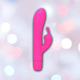 The B Swish Limited Edition Bwild Infinite Bunny - Sunset Pink, a bright pink silicone clitoral stimulator with a curved shaft and smaller extension, is shown against a soft pastel background of pink, white, and blue circles.