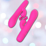 Two B Swish Limited Edition Bwild Infinite Bunny - Sunset Pink silicone dual-prong clitoral stimulators appear against a softly blurred background with white and pastel bokeh lights.