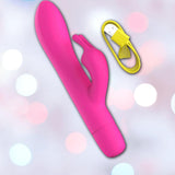 The B Swish Limited Edition Bwild Infinite Bunny - Sunset Pink, a dual-ended silicone stimulator with a rabbit-shaped tip, is displayed on a pastel bokeh background next to its yellow USB charging cable.