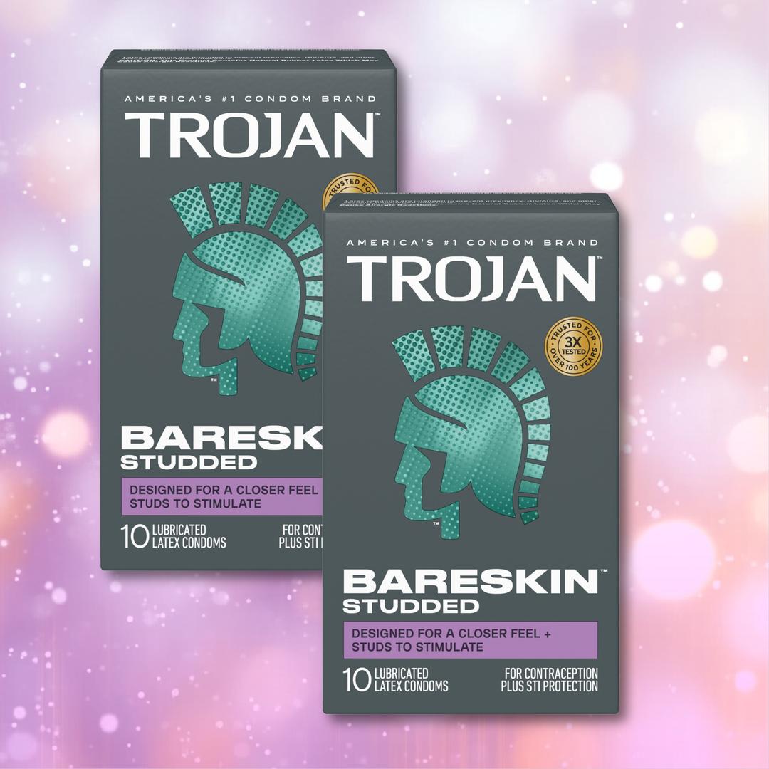 Two boxes of Trojan Studded Bareskin Condoms from Trojan, each featuring a teal Roman helmet design, are showcased against a vibrant pink, purple, and yellow bokeh background. These ultra-thin textured condoms offer heightened sensation along with dependable protection, with every box containing 10 lubricated latex condoms.