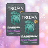 Two boxes of Trojan Studded Bareskin Condoms from Trojan, each featuring a teal Roman helmet design, are showcased against a vibrant pink, purple, and yellow bokeh background. These ultra-thin textured condoms offer heightened sensation along with dependable protection, with every box containing 10 lubricated latex condoms.