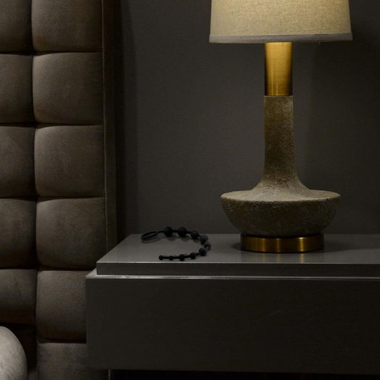 In a dimly lit room, a textured bedside lamp sits on a sleek dark nightstand with Blush Novelties' Anal Adventures Platinum Ultrasilk Silicone 10 Beads nearby. The cushioned dark wall panels enhance the cozy atmosphere, perfect for intimate moments. 1080