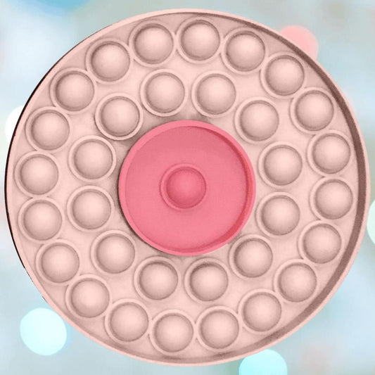 The Kheper Games Boob Pop-It Toy is a round, pastel stress relief toy with a pink center and two bubble rings, resembling bubble wrap. It's shown against a softly blurred blue and pink background. 1080