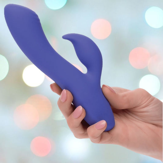 A hand holds the CalExotics Connect Dual Stimulator, a blue silicone vibrator with dual arms, set against a backdrop of pastel bokeh lights. 1080