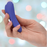 A hand with pink nails holds the small, blue, oval-shaped CalExotics Connect Panty Teaser, featuring a textured grip for app-controlled pleasure. The softly blurred background with pastel-colored bokeh lights evokes a sense of playful intimacy.