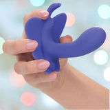 A hand holds the CalExotics Connect Venus Butterfly App Remote Control Dual Stimulator, a blue silicone device featuring a distinctive wing-like design. With dual motors for enhanced stimulation, it is set against a background of soft, blurry circular lights in pastel hues.